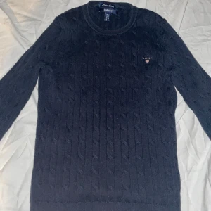 Mörkblå stickad tröja från GANT - Snygg mörkblå stickad tröja från GANT med klassiskt kabelstickat mönster och rund hals. Tröjan har långärmade och en liten broderad logga på bröstet. Perfekt för dig som gillar stilrena och tidlösa plagg.