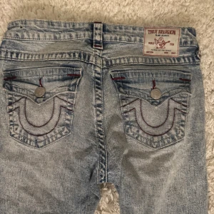 True Religion ljusa bootcut jeans - Säljer ett par ljusblå jeans från True Religion med bootcut passform och slitna detaljer på benen. Jeansen har röda sömmar, klassiska fickor med lock och silverfärgade knappar. Materialet är jeans och midjan är midwaist. Perfekt för dig som gillar street och retrovibe.