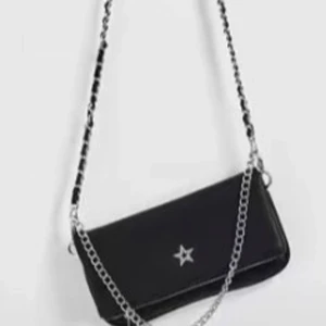 Svart axelväska med kedja och stjärna - Cool svart axelväska med silverfärgad kedjerem och en liten stjärndetalj framtill. Väskan har en stilren, rektangulär form och passar perfekt för dig som gillar en edgy look. Materialet ser ut som skinn och kedjan ger en extra touch.