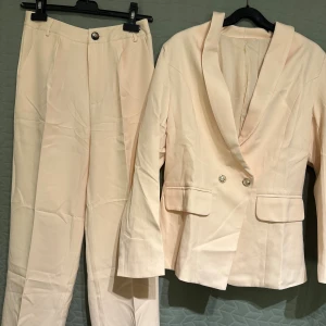 Aprikosfärgad kostym  - Damkostym i ljus aprikosfärg med klassisk kavaj och raka byxor. Kavajen har dekorativa guldknappar både fram och vid ärmslut. Jätteskönt material. ❕Helt ny. Aldrig använd. Kvar i originalförpackning❕ Kavaj - Längd: ca 68cm, Byst (omkring): ca 96cm, Midja (omkring): ca 96cm. Byxor - Längd: ca 100cm, Midja (omkring): ca 72cm, Benslut (omkring): ca 44cm. 