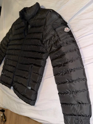 Svart Moncler pufferjacka herr - Snygg svart pufferjacka från Moncler med klassisk logga på ärmen. Jackan har ståkrage, två sidofickor med dragkedja och quiltad design. Perfekt för dig som vill ha en stilren och varm jacka med grym passform.