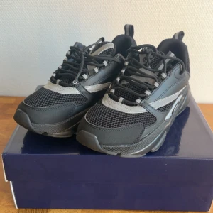 Dior B22 - Chunky svarta Dior sneakers med grå detaljer, tillverkade i mesh och skinn. Snyggt sportig design med platt sula, snörning och rund tå. Märkeslogga på sidan och coola paneler som ger en modern vibe. Perfekt för dig som gillar statement-sneakers.