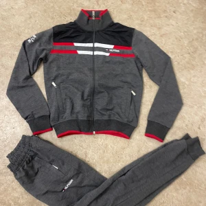 Grå tracksuit från Tommy Hilfiger - Snygg grå tracksuit från Tommy Hilfiger med röda och vita detaljer. Jackan har hel dragkedja, ståkrage och svarta paneler upptill. Byxorna har resår i midjan och logotyp på benet. Perfekt för chill eller sportiga dagar.