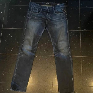 Anvbass replay nytt skick - Säljer ett par Replay Anbass jeans i mörkblå tvätt med snygga slitningar och klassisk femficksdesign. Jeansen har raka ben och normal passform, med Replay-läderpatch bak i midjan. Perfekta för dig som gillar stilrena och bekväma jeans.
