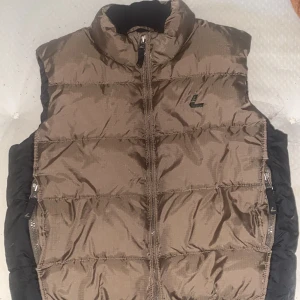 Brun och svart dunväst med dragkedja - Snygg dunväst i brunt och svart med hög krage och dragkedja framtill. Västen har quiltad design och två sidofickor med dragkedja. Perfekt för lager-på-lager och chill stil. Diskret L-logga på bröstet.