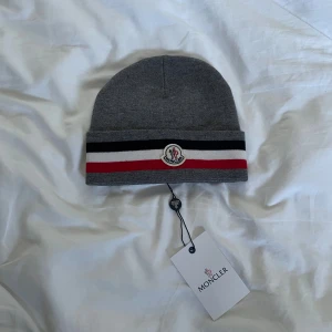 Grå M mössa med ränder - Snygg grå mössa från M med klassiska ränder i svart, vitt och rött på uppviket. Märkets logga är broderad framtill. Mössan är stickad och har en mjuk känsla, perfekt för kyliga dagar. Taggen sitter kvar.