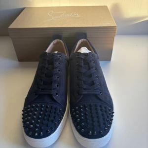 Christian Louboutin sneakers blå med nitar - Snygga mörkblå sneakers från Christian Louboutin med klassisk röd sula och coola svarta nitar på tån. Skorna har snörning och är tillverkade i mocka med vit platt sula. Helt nya aldrig använda st44