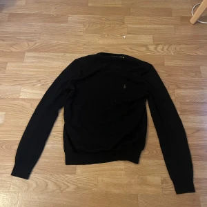 Svart merinoulltröja från Polo Ralph Lauren - Slim fit svart tröja från Polo Ralph Lauren i mjuk och tvättbar merinoull. Klassisk design med rund hals och diskret broderad logga på bröstet. Långärmad och ribbade muddar vid ärmslut och nederkant. Perfekt för dig som gillar stilrena och tidlösa plagg.