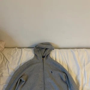Grå hoodie från Polo Ralph Lauren - Säljer en grå hoodie från Polo Ralph Lauren i storlek L. Tröjan har huva, dragkedja framtill och ett litet blått Polo-logo broderat på bröstet. Perfekt för chill dagar och enkel att matcha med jeans eller joggers.