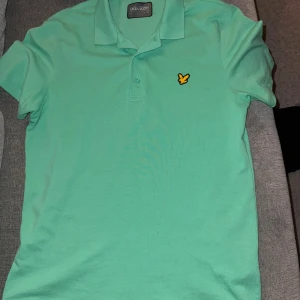 Mintgrön pikétröja från Lyle & Scott - Fräsch mintgrön pikétröja från Lyle & Scott med klassisk krage och två knappar framtill. Tröjan har korta ärmar och den ikoniska gula fågeln broderad på bröstet. Perfekt för dig som gillar stilrena och sportiga plagg.