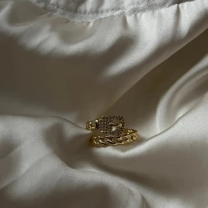 Guldfärgad ring med kedjedetalj - Snygg guldfärgad ring med kedjeinspirerad design och ett spänne framtill som är dekorerat med små glittrande stenar. Ringen har en chunky känsla och passar perfekt för dig som gillar statement-smycken med coola detaljer. 17mm går att justera🩷 18k guldpläterad 