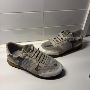 Valentino Rockrunner  - Snygga Valentino Rockrunner sneakers i beige och grått med coolt camouflagemönster. Skorna har mesh och mocka-paneler, vita detaljer vid tån och hälen samt snörning. Gummisula med nitar på hälen ger extra edge. Perfekta för dig som gillar streetstyle.
