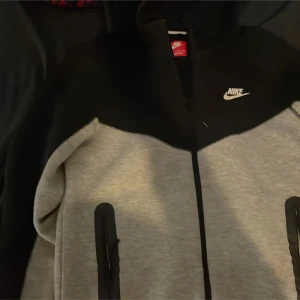 Nike tech svart grå  - Nike vindjacka i svart och ljusgrått med vit logga på bröstet. Jackan har hel dragkedja framtill och två svarta dragkedjeförsedda fickor. Sportig design med mjukt material som passar perfekt till streetwear. Snygg kontrast mellan färgerna och stilren look.