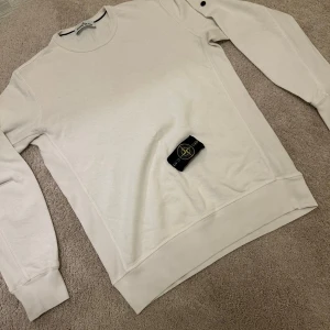 Beige sweatshirt från Stone Island - Säljer en clean vitt sweatshirt från Stone Island med klassisk rund hals och ribbade muddar. Tröjan har den ikoniska avtagbara logotypen på ärmen och är gjord i mjuk bomull. Perfekt för dig som gillar stilrena och exklusiva plagg.