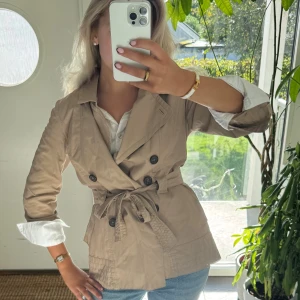 Beige trenchcoat med bälte och knappar - Snygg beige trenchcoat med dubbelknäppning och bälte i midjan. Jackan har klassisk krage, svarta knappar och långa ärmar som kan vikas upp för en chill look. Perfekt för dig som gillar stilrena och tidlösa plagg. Passar S-M