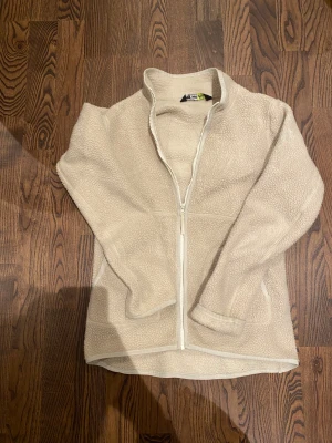 Beige fleecejacka från Helly Hansen - Mjuk och varm beige fleecejacka från Helly Hansen med hel dragkedja framtill och ståkrage. Jackan har två sidofickor och HH-logga broderad på ärmen. Perfekt för kyliga dagar och enkel att slänga på sig när du vill vara bekväm.