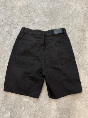 Svarta denimshorts från Stay - Svarta denimshorts från Stay med klassisk femficksdesign och bälteshällor. Modellen är rak och lite längre, med hög midja och knapp- samt dragkedjestängning. Perfekta för en clean och stilren look.