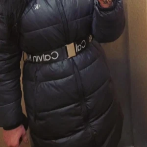 Svart lång pufferjacka Calvin Klein - Säljer en svart lång pufferjacka från Calvin Klein med coolt logobälte i midjan. Jackan har dragkedja framtill, hög krage och långa ärmar. Perfekt för kalla dagar när du vill vara både varm och snygg.