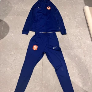 Nike fotboll tracksuit - Säljer en blå hoodie från Nike med dragkedja och huva. Tröjan har ett vitt Nike-märke och ett orange lejonemblem på bröstet. Tillverkad i polyester och har långa ärmar samt fickor med dragkedja. Perfekt för träning eller sportiga dagar.