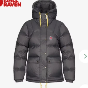Mörkgrå dunjacka från Fjällräven - Snygg mörkgrå dunjacka från Fjällräven med stor huva och gula snören. Jackan har två stora fickor med knappar, Fjällräven-logga på bröstet och dragkedja med knappslå. Perfekt för kalla dagar och har en riktigt puffig look.