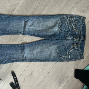 Jeans - Snygga blå jeans från Morgan köpta i Spanien med utsvängda bootcut passform och slitna detaljer. Jeansen har dekorativa bakfickor med broderade mönster och knappar, samt klassisk femficksdesign. Materialet är jeans med kontrastsömmar och cool tvättad sliten look. Väldigt lågmidjade storlek S-M. 