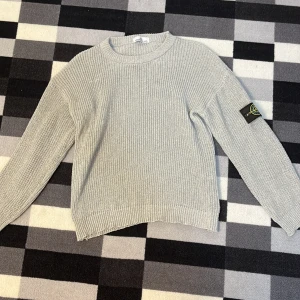 Ljusgrå stickad Stone Island tröja - Snygg ljusgrå stickad tröja från Stone Island med klassisk patch på vänster ärm. Tröjan har rund hals, ribbade muddar och är långärmad. Perfekt för dig som gillar clean och stilren design med street-vibe.