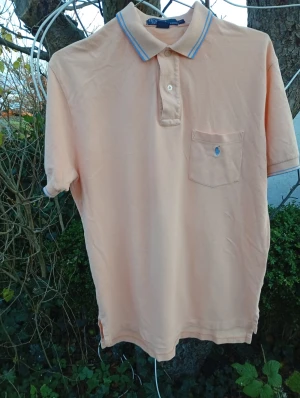 Beige pikétröja från Polo Ralph Lauren - Snygg beige pikétröja från Polo Ralph Lauren med klassisk krage och blå ränder. Tröjan har korta ärmar, knappslå och en bröstficka med blå broderad logga. Tillverkad i mjuk bomull som är skön mot huden. Perfekt för dig som gillar stilren och enkel design.