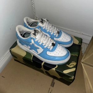 BAPE STA sneakers blå och vita - Säljer ett par BAPE STA sneakers i blått och vitt med klassisk stjärnlogga på sidan. Skorna har snörning, rund tå och platt sula. Ovandelen är i blankt skinn och de kommer med originalkartong i camomönster. Perfekt för dig som gillar streetwear och vill sticka ut.