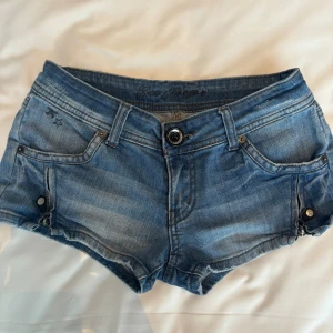 Blå jeansshorts från Coco Bongo - Snygga blå jeansshorts från Coco Bongo med låg midja och klassisk femficksdesign. Shortsen har slitna detaljer, knappstängning och coola små stjärnor på ena fickan. Perfekta för varma dagar och har en riktigt chill vibe.