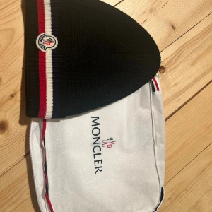 Svart Moncler mössa - Svart stickad mössa från Moncler med klassiska ränder i vitt, rött och marinblått längs kanten. Mössan har Moncler-logga framtill och är tillverkad i mjukt material som sitter skönt på huvudet. Perfekt för dig som vill ha en snygg och stilren accessoar. Använd knappt en vinter, kvitto finns! Samt följer etikett och brev från moncler😃