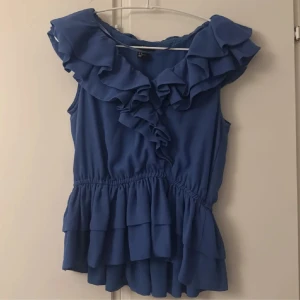 Blå volangtopp med ärmlös design - Supersöt blå topp med volanger längs halsringningen och nertill. Modellen är ärmlös och har en markerad midja med resår för en snygg siluett. Perfekt för dig som gillar feminina detaljer och en snygg känsla. 💞🪽