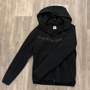 Svart Nike tech fleece tröja  - Svart Nike tech fleece tröja med hel dragkedja framtill. Tröjan har en diskret Nike-logga på bröstet och en snygg detalj med ett horisontellt band över bröstet. Perfekt för chill dagar eller när du vill ha en sportig look. Tröjan är i storlek S, priset går självklart att diskuteras. Nypris ca 1000 kr.