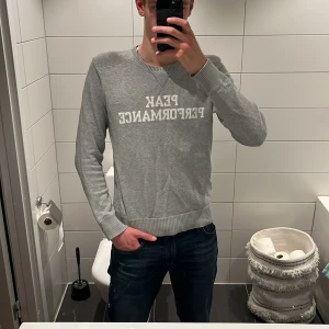Grå stickad tröja Peak Performance - Snygg grå stickad tröja från Peak Performance med vit logotyp framtill. Tröjan har rund hals, ribbade muddar och långärmad passform. Perfekt för dig som gillar stilrena och klassiska plagg. Jag på bilden är 187cm, tröjan kostar 1299 kr ny, tveka inte på att höra av er om prisförslag och frågor 
