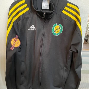 Svart Adidas vindjacka med gula ränder - Svart träningsjacka från Adidas med gula ränder längs ärmarna, dragkedja framtill och logga på bröstet. Jackan har tryck från IK Sävehof och Hard Rock Cafe på ena ärmen samt Stadium-tryck på ryggen. Stl 152, 11-12 år 