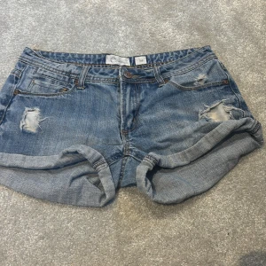 Blå jeansshorts från Cubus, strl 34 - Säljer ett par blå jeansshorts från Cubus i storlek 34. De har slitna detaljer, upprullade benslut och klassisk femficksdesign. Perfekta för sommardagar och ger en avslappnad vibe. Materialet är bomull och shortsen har normal passform.