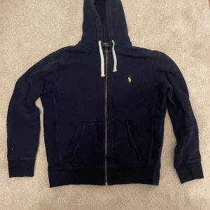 Marinblå hoodie från Polo Ralph Lauren - Snygg marinblå hoodie från Polo Ralph Lauren med gul broderad logga på bröstet. Tröjan har huva med vita snören, dragkedja framtill och två fickor. Tillverkad i mjuk bomull, perfekt för chill dagar. Klassisk och stilren design. Har en skada på armbågen. 