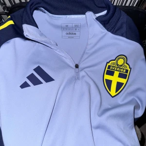 Svenska landslaget Adidas half zip tröja - Säljer en snygg ljusblå och marinblå långärmad tröja från Adidas med Sveriges landslagsemblem på bröstet. Tröjan har half zip framtill, gula detaljer på axlarna och är i slim fit. Perfekt för fotbollsträning eller när du vill representera Sverige.