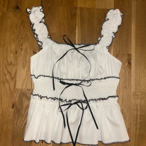 Vit volangtopp med svarta detaljer  - Supersöt vit topp från Shein med volangprydda axelband och svarta kontrastsömmar. Smockad midja och tre svarta knytband framtill ger en extra touch. 