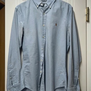 Blå skjorta från Polo Ralph Lauren - Snygg ljusblå skjorta från Polo Ralph Lauren i slim fit-modell. Klassisk button-down krage, broderad logga på bröstet och knappar hela vägen. Tillverkad i mjuk bomull, perfekt för dig som gillar stilrena och tidlösa plagg.