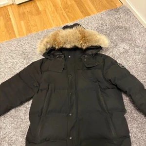 Canada goose Wyndham  - Säljer en svart dunjacka från Canada Goose med stor huva och fluffig pälsdetalj. Jackan har flera fickor, knappar och dragkedja framtill. Perfekt för kalla vinterdagar och har en snygg, klassisk look med black lable patch på ärmen.   L men passar till M också