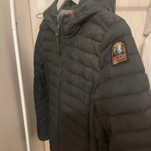 Svart dunjacka från Parajumpers S - Snygg svart dunjacka från Parajumpers i storlek S. Jackan har huva, dragkedja framtill och ett coolt patch på ärmen. Lättviktig och quiltad design som ger en schysst siluett. Perfekt för dig som vill ha en stilren och funktionell jacka med streetkänsla.