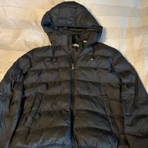 Svart pufferjacka från GANT - Svart pufferjacka från GANT med huva och dragkedja framtill. Jackan har quiltad design, två sidofickor och GANT-logga med emblem på bröstet. Perfekt för kalla dagar och har en snygg, klassisk look som funkar till det mesta.