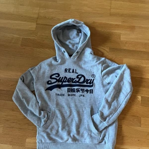 Grå Superdry hoodie med blått tryck - Snygg grå hoodie från Superdry med stort blått broderat logotryck på bröstet och japanska tecken. Tröjan har huva med metallöglor, känguruficka och ribbade muddar. Perfekt för chill dagar och streetstyle. Pris därav defekt vid luvan annars bra skick, hör av dig gällande frågor😎 storlek M 