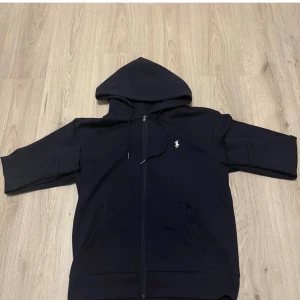 Svart hoodie från Polo Ralph Lauren - Svart hoodie från Polo Ralph Lauren med klassisk broderad logga på bröstet. Tröjan har huva med snörning, hel dragkedja och två fickor framtill. Materialet är mjuk bomull och passformen är normal. Perfekt för chill dagar och streetwear.
