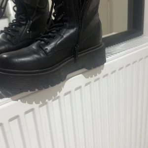 Svarta boots med platå och snörning - Säljer ett par svarta boots med grov platåsula och snörning framtill. Skorna har dragkedja på sidan för enkel på- och avtagning. Tillverkade i skinnimitation med rund tå och robust design som ger en cool vibe till din outfit.