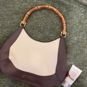 Axelväska med bambuhandtag - Snygg axelväska i gott skick och med bambuhandtag. Väskan är vit och brun och small i modellen. Insidan är fodrad med mönstrat tyg och magetlås. Finns även axelrem som tillkommer. Köpt i en second hand i Kyoto, säljer då den inte kommer till användning.