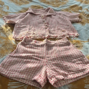 Rutig tweedshorts i rosa och vit - Säljer ett par snygga rutig tweedshorts i rosa och vit med hög midja. Materialet är grovt vävt och har en lyxig känsla, perfekt för dig som gillar att sticka ut. Shortsen har en klassisk form och matchar toppen med fransiga detaljer.