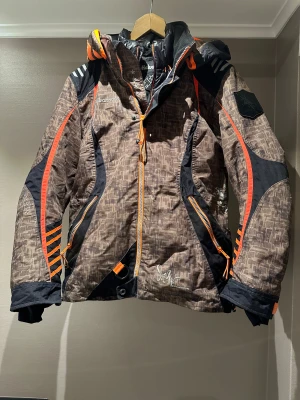 Halvarssons brun och svart skoter dress  - Säljer en brun och svart skoter dress  från Halvarssons med orange detaljer och grafiskt mönster. Jackan har huva, dragkedjor. På dressen är det en över jacka och sen en innerjacka som är lös tagbar sen på byxorna så är det ett snö lås i midjan och nere vid fotlederna 