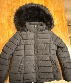 Svart pufferjacka med fuskpäls från Navahoo - Säljer en svart pufferjacka från Navahoo i storlek XL. Jackan har en mysig huva med fuskpäls, quiltad design och både dragkedja och knappar framtill. Insidan är fodrad med mjuk, svart fleece för extra värme. Perfekt för kalla dagar! Passar även till storlek L 