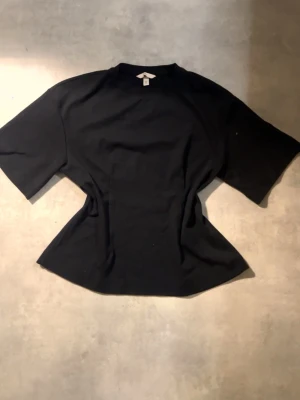 Svart kortärmad topp från HM - Svart topp från HM med korta ärmar och rund hals. Modellen har en lätt figurnära passform med markerad midja och lite utsvängd nederkant. Materialet är mjuk bomull som känns skönt mot huden. Perfekt att styla med jeans eller kjol för en clean look.